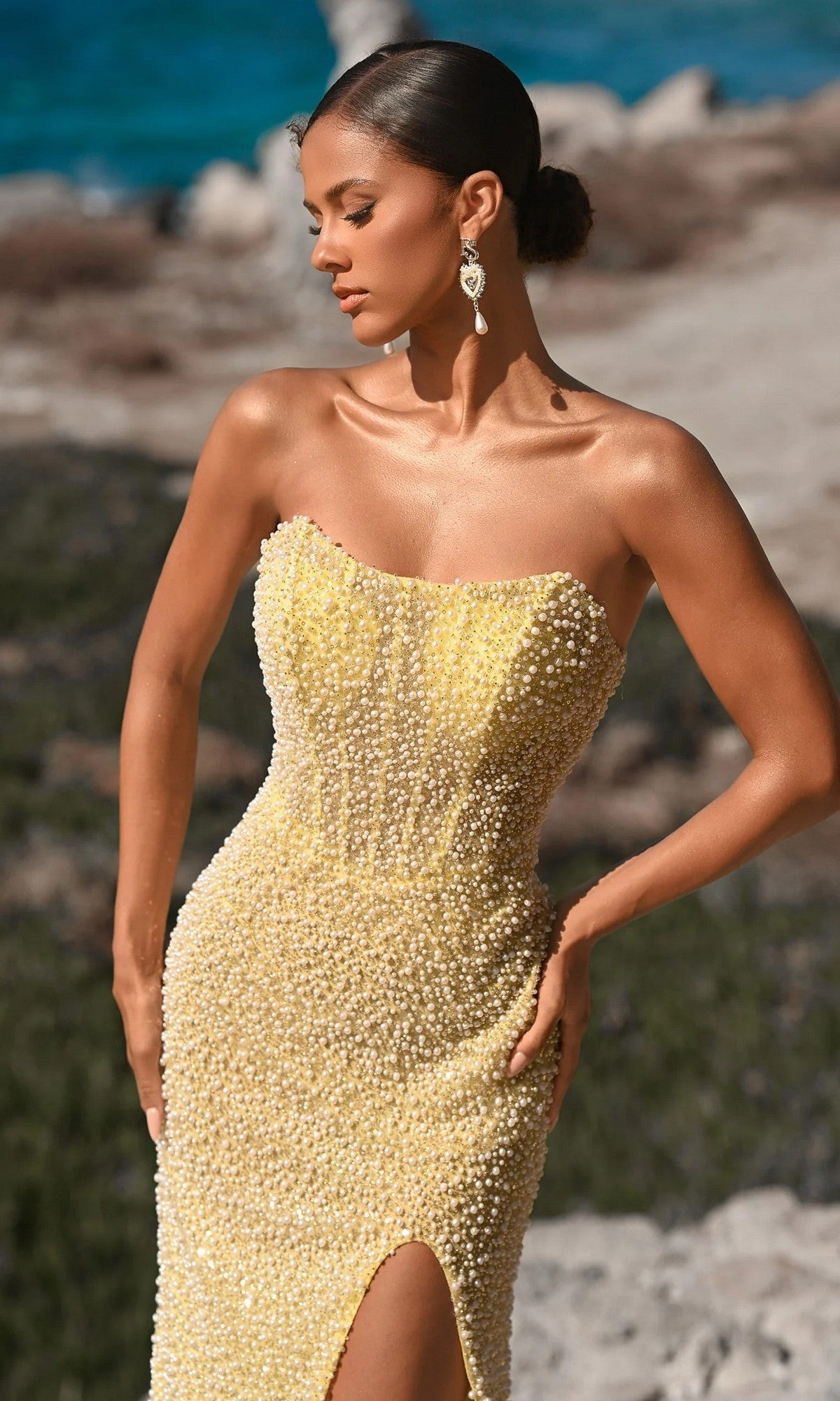 A woman in the yellow Portia and Scarlett PS26173 long formal dress with beaded details, a sheer corset bodice, strapless neckline, and thigh-high slit stands outdoors by rocky terrain and the ocean.